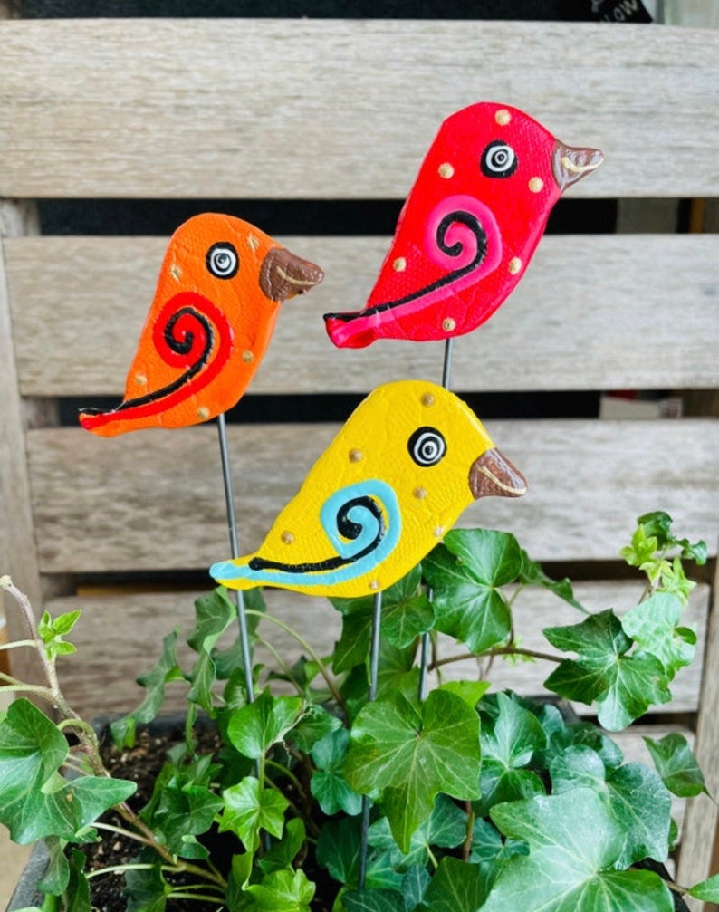 May include: Three colorful ceramic bird figurines with painted details, each perched on a metal stake, are displayed in a potted plant. The birds are red, orange, and yellow with black and blue accents.