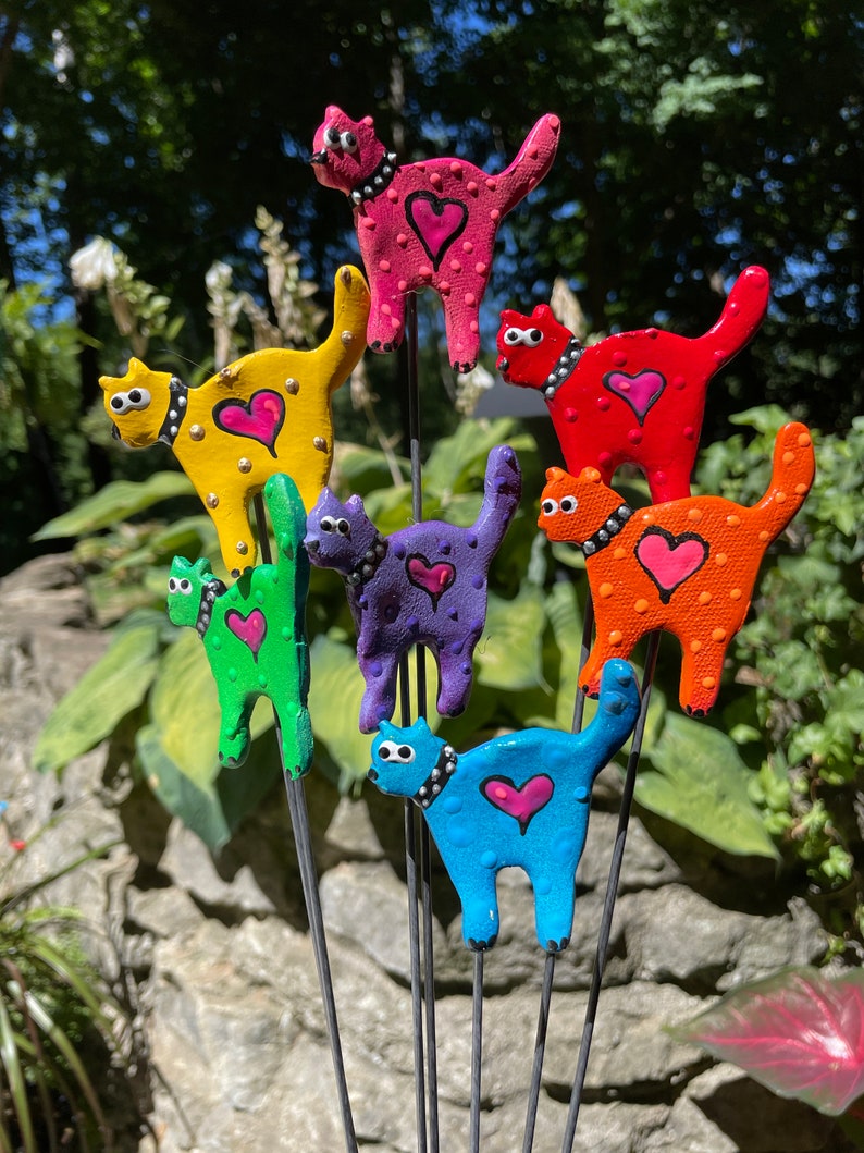 Puede incluir: Siete figuritas de cer&aacute;mica de gatos coloridos con corazones en el pecho, cada una sobre una estaca de metal negro. Los gatos est&aacute;n pintados en tonos de rosa, amarillo, verde, morado, naranja y azul.