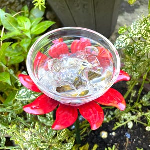 May include: A red metal flower with a glass bowl in the center filled with ice cubes. The flower is sitting in a pot of green plants.