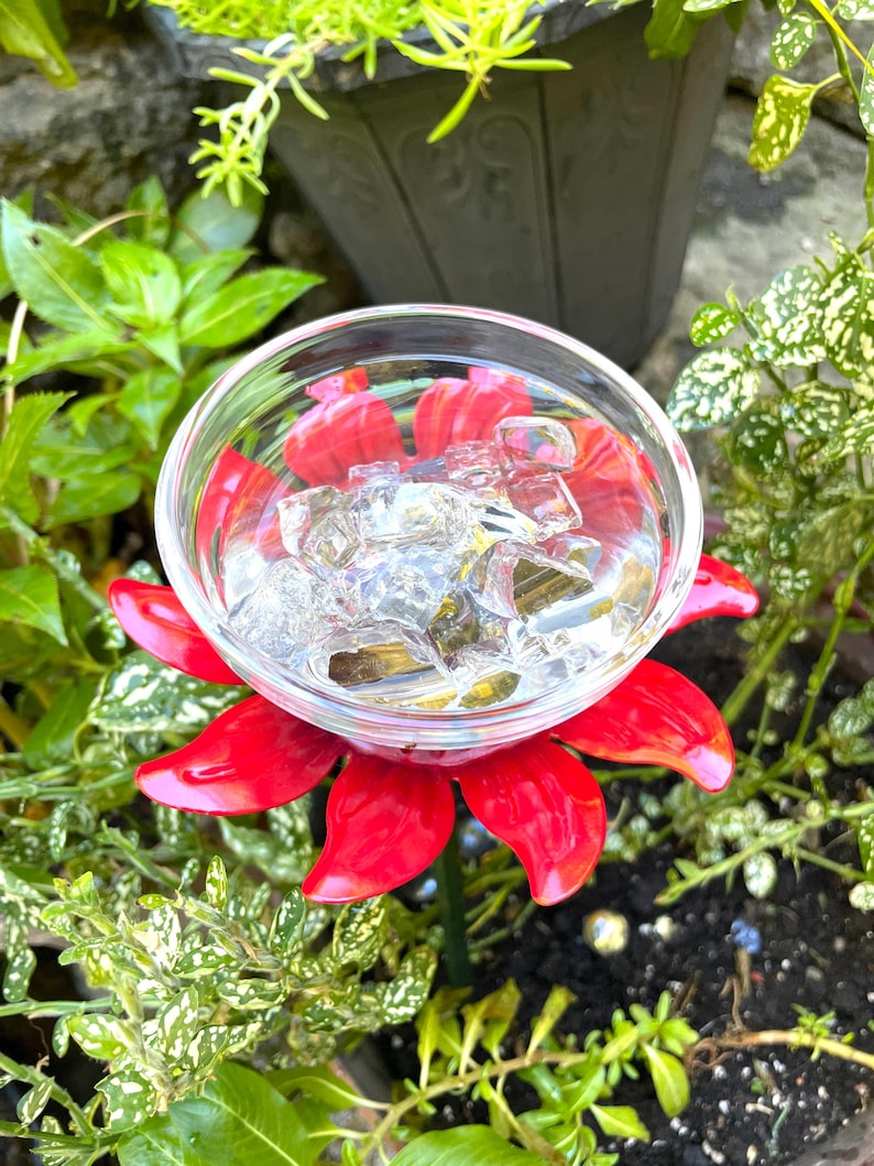 May include: A red metal flower with a glass bowl in the center filled with ice cubes. The flower is sitting in a pot of green plants.