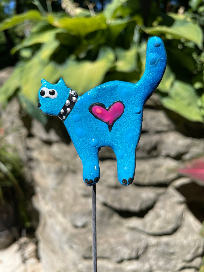 Puede incluir: Una figurita de cer&aacute;mica azul de un gato con lunares blancos y un coraz&oacute;n rosa en la espalda. El gato tiene ojos negros y un collar negro con tachuelas. La figurita est&aacute; unida a una estaca de metal.