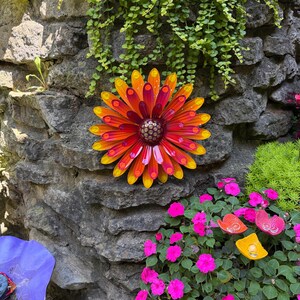 May include: A circular metal wall art piece shaped like a flower with multiple layers of pointed petals in vibrant shades of yellow, orange, and deep red. The center of the flower features a textured, dark brown dome with small raised dots.