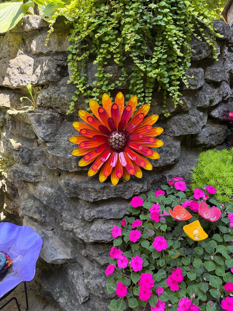 May include: A circular metal wall art piece shaped like a flower with multiple layers of pointed petals in vibrant shades of yellow, orange, and deep red. The center of the flower features a textured, dark brown dome with small raised dots.