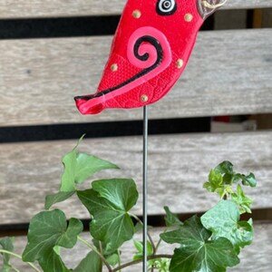 May include: A red ceramic bird with black and pink details and gold accents. The bird is attached to a metal stake.