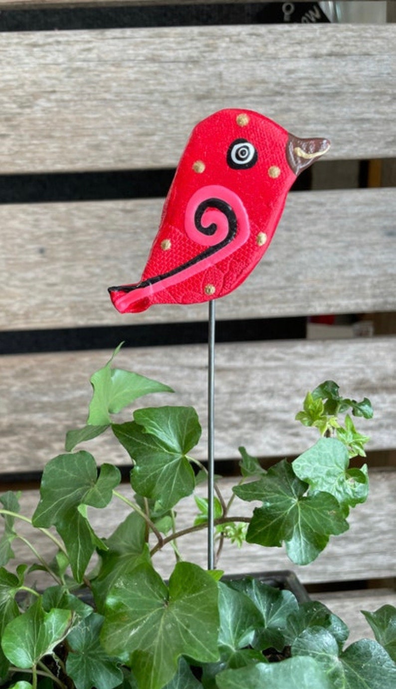 May include: A red ceramic bird with black and pink details and gold accents. The bird is attached to a metal stake.
