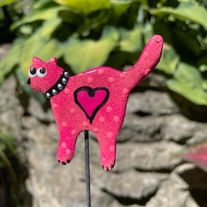 Puede incluir: Figura de cer&aacute;mica de gato rosa con lunares blancos y un coraz&oacute;n rosa en un lado. El gato tiene ojos negros y un collar negro con tachuelas plateadas. La figura est&aacute; unida a una estaca de metal negro.