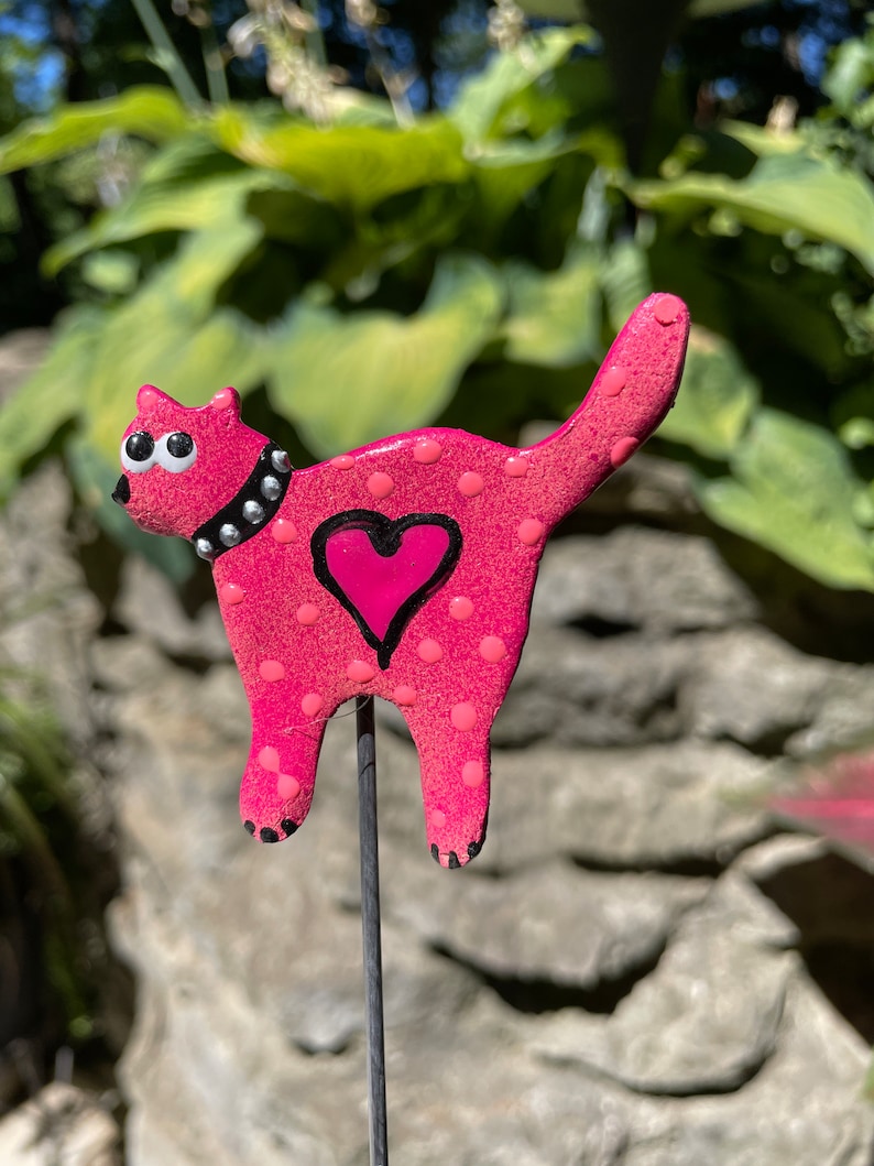 Puede incluir: Figura de cer&aacute;mica de gato rosa con lunares blancos y un coraz&oacute;n rosa en un lado. El gato tiene ojos negros y un collar negro con tachuelas plateadas. La figura est&aacute; unida a una estaca de metal negro.