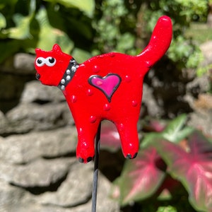 Puede incluir: Figura de gato de cer&aacute;mica roja con lunares blancos, un collar negro tachonado y un coraz&oacute;n rosa en el pecho. El gato est&aacute; de pie sobre una estaca de metal negro.
