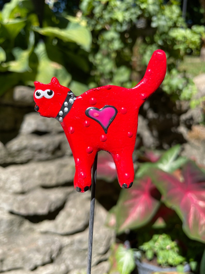 Puede incluir: Figura de gato de cer&aacute;mica roja con lunares blancos, un collar negro tachonado y un coraz&oacute;n rosa en el pecho. El gato est&aacute; de pie sobre una estaca de metal negro.