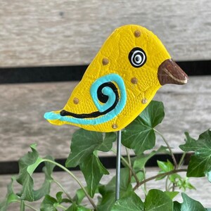May include: A yellow ceramic bird with blue and black painted details, perched on a metal stake. The bird has a textured surface and a black eye with a white circle in the center.