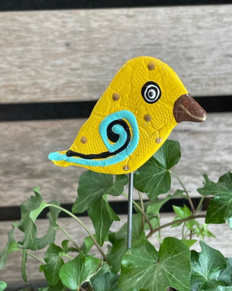 May include: A yellow ceramic bird with blue and black painted details, perched on a metal stake. The bird has a textured surface and a black eye with a white circle in the center.