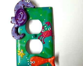 May include: A rectangular electrical outlet wall plate with a green background featuring painted sea life. The plate includes a purple seahorse, a pink crab, a yellow starfish, and an orange fish, all with raised textures and white googly eyes.