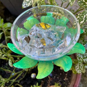 May include: A green metal flower with a clear glass bowl filled with ice cubes. The flower is sitting in a pot of dirt with green plants.