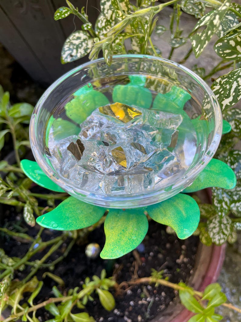 May include: A green metal flower with a clear glass bowl filled with ice cubes. The flower is sitting in a pot of dirt with green plants.