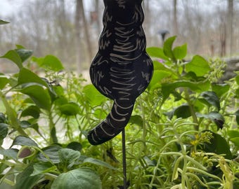 May include: Black and white ceramic cat figurine garden stake. The cat is sitting with its back to the viewer and its tail curled around its legs. The stake is inserted into a pot of green plants.