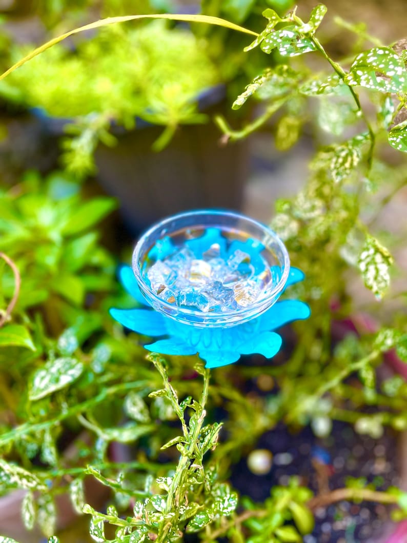 May include: A blue plastic flower with a clear glass bowl in the center. The bowl is filled with clear ice cubes. The flower is sitting on a bed of green leaves.