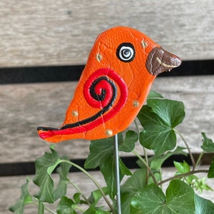 May include: Orange and red clay bird with black and gold accents. The bird has a swirl design on its body and a brown beak. The bird is attached to a metal stake.