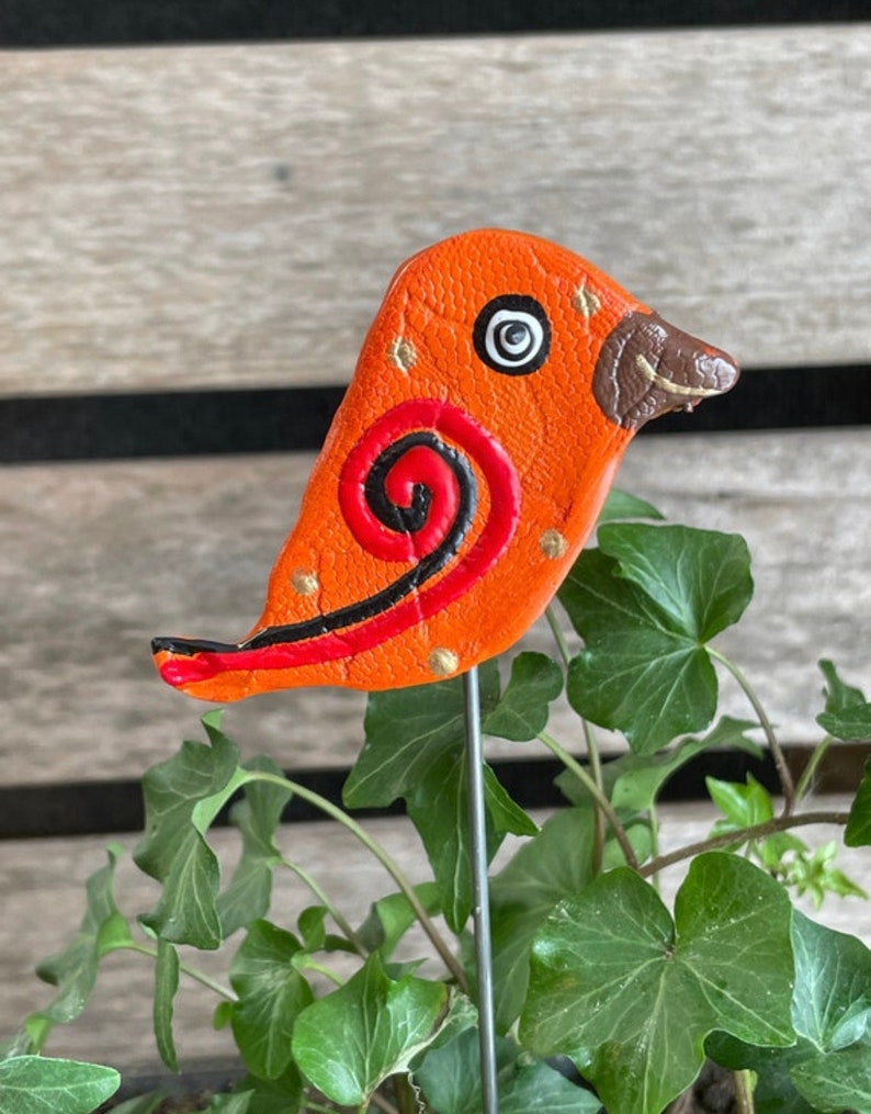May include: Orange and red clay bird with black and gold accents. The bird has a swirl design on its body and a brown beak. The bird is attached to a metal stake.