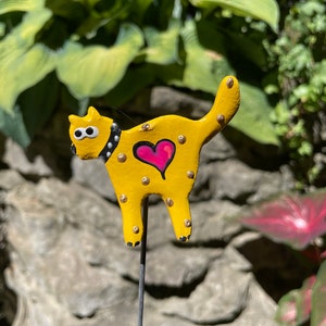 Puede incluir: Una figurita de cer&aacute;mica de gato amarillo con ojos negros, un collar blanco y negro y un coraz&oacute;n rosa en el pecho. El gato est&aacute; de pie sobre una estaca de metal negro.