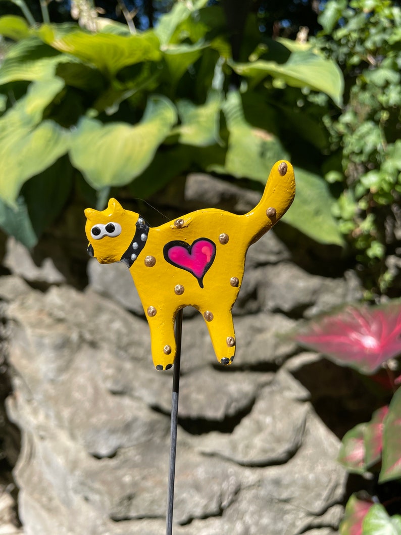 Puede incluir: Una figurita de cer&aacute;mica de gato amarillo con ojos negros, un collar blanco y negro y un coraz&oacute;n rosa en el pecho. El gato est&aacute; de pie sobre una estaca de metal negro.