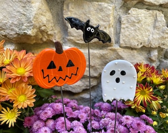 May include: Three Halloween-themed garden stakes: a black bat with white eyes, an orange pumpkin with a black jack-o'-lantern face, and a white ghost with black eyes and a mouth.