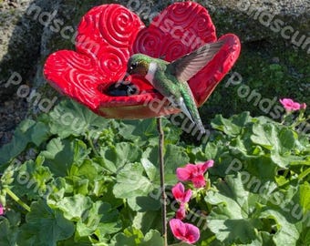 Op de afbeelding: Een rode, bloemvormige kolibrie feeder met een kolibrie erop. De feeder is gemaakt van een glanzend rood materiaal en heeft een decoratief wervelpatroon. De kolibrie heeft groene en witte veren. De feeder staat in een tuin.
