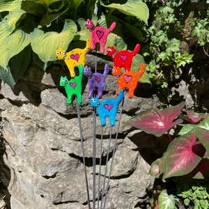 Puede incluir: Un conjunto de ocho estacas de jard&iacute;n de gato de colores. Cada estaca presenta un gato de color diferente con un coraz&oacute;n rosa en el pecho. Los gatos est&aacute;n dispuestos en un patr&oacute;n de arco&iacute;ris, con el gato rojo en la parte superior y el gato azul en la parte inferior.