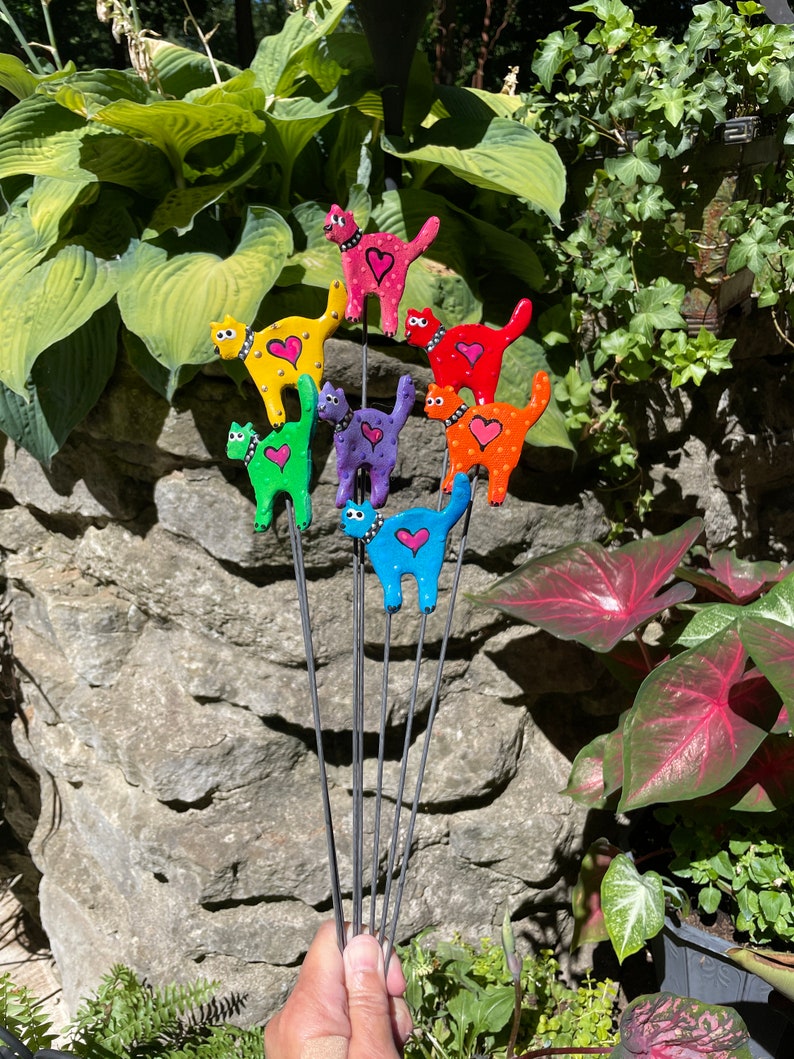 Puede incluir: Un conjunto de ocho estacas de jard&iacute;n de gato de colores. Cada estaca presenta un gato de color diferente con un coraz&oacute;n rosa en el pecho. Los gatos est&aacute;n dispuestos en un patr&oacute;n de arco&iacute;ris, con el gato rojo en la parte superior y el gato azul en la parte inferior.