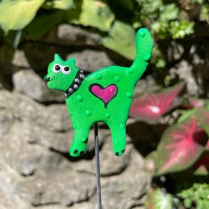 Puede incluir: Una figurita de gato verde con un coraz&oacute;n rosa y un collar negro con tachuelas. El gato est&aacute; de pie sobre una estaca de metal.