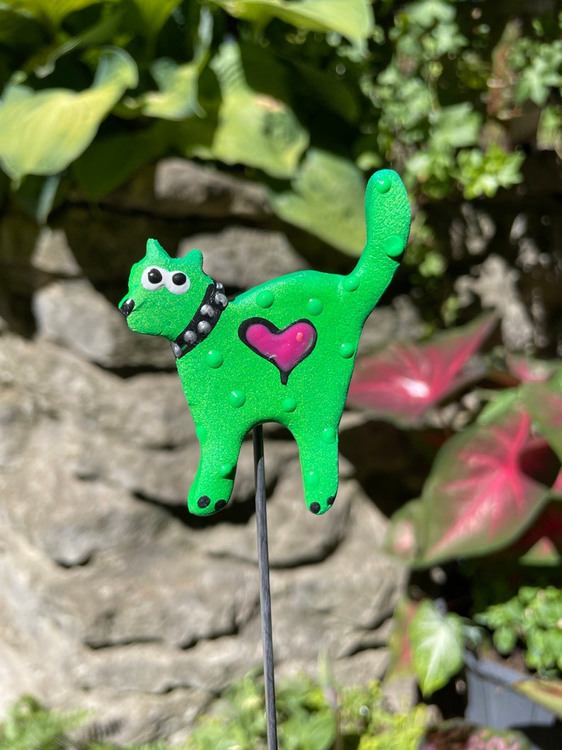 Puede incluir: Una figurita de gato verde con un coraz&oacute;n rosa y un collar negro con tachuelas. El gato est&aacute; de pie sobre una estaca de metal.