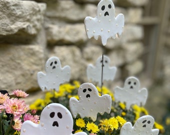 May include: A group of six white ceramic ghost figurines with black eyes and mouths are placed in a pot of pink and yellow flowers. The ghosts are decorated with white dots and have a dripping effect.