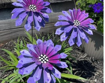 May include: Three purple metal flowers with white polka dots in the center. The flowers are on black metal stems and are in a garden setting.