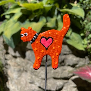 Puede incluir: Figurita de gato de cer&aacute;mica naranja con lunares blancos y un coraz&oacute;n rosa en la espalda. El gato lleva un collar negro con tachuelas plateadas y tiene ojos blancos. La figurita est&aacute; unida a una estaca de metal.