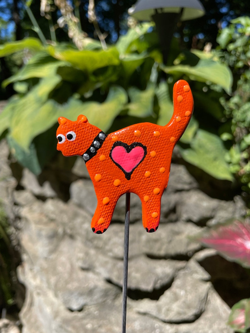 Puede incluir: Figurita de gato de cer&aacute;mica naranja con lunares blancos y un coraz&oacute;n rosa en la espalda. El gato lleva un collar negro con tachuelas plateadas y tiene ojos blancos. La figurita est&aacute; unida a una estaca de metal.