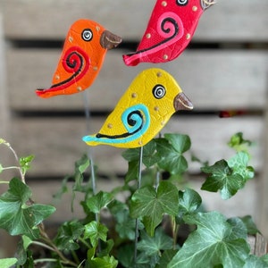 May include: Three colorful ceramic bird figurines with painted details, including swirls and dots, are attached to metal stakes. The birds are red, orange, and yellow and are arranged in a pot of green ivy.