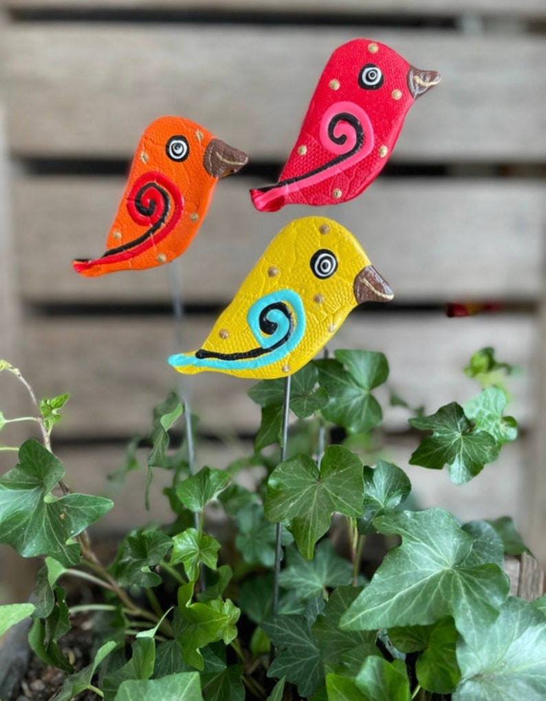 May include: Three colorful ceramic bird figurines with painted details, including swirls and dots, are attached to metal stakes. The birds are red, orange, and yellow and are arranged in a pot of green ivy.
