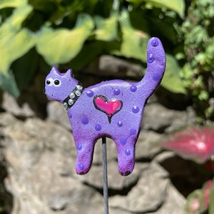 Puede incluir: Figura de cer&aacute;mica de gato p&uacute;rpura con ojos blancos, un coraz&oacute;n rosa y lunares blancos. El gato est&aacute; de pie sobre una estaca de metal.