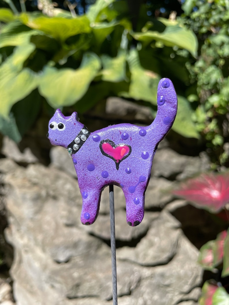Puede incluir: Figura de cer&aacute;mica de gato p&uacute;rpura con ojos blancos, un coraz&oacute;n rosa y lunares blancos. El gato est&aacute; de pie sobre una estaca de metal.