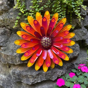 May include: A multi-layered metal flower wall decoration featuring elongated petals in a gradient of yellow, orange, and magenta. The center is a dark purple dome with a pattern of raised gold-colored dots, and each petal tip has a small yellow dot.