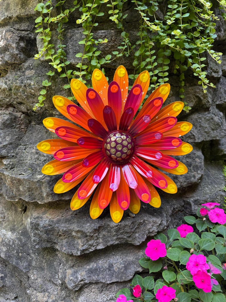 May include: A multi-layered metal flower wall decoration featuring elongated petals in a gradient of yellow, orange, and magenta. The center is a dark purple dome with a pattern of raised gold-colored dots, and each petal tip has a small yellow dot.