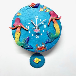 May include: A decorative wall clock with a blue face, adorned with two mermaids with pink and yellow hair and tails. The clock features white hands, a pink fish, and a crab. Small yellow fish and a yellow fish pendulum complete the design.