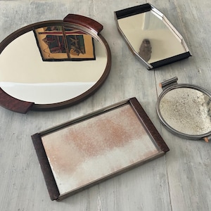 May include: A collection of four serving trays including a large round wooden-handled tray, a rectangular tray with black handles, a smaller round metal tray with side handles, and a rectangular tray with dark wooden ends and a weathered surface.