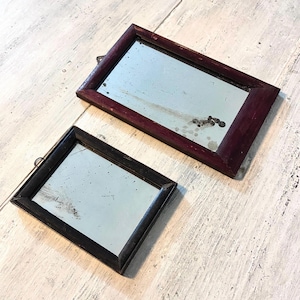 Puede incluir: Dos espejos rectangulares antiguos con marcos de madera oscura. Uno tiene un marco burdeos oscuro y el otro un marco negro. Ambos espejos tienen un pequeño lazo de metal en la parte superior para colgar.