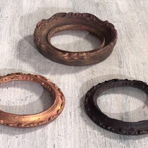 May include: Three decorative, oval-shaped frames in varying shades of brown. The top frame is a dark, textured brown, while the bottom left frame has a metallic copper finish. The bottom right frame is a dark, matte brown.