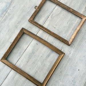 May include: Two rectangular, vintage-style picture frames with a warm, golden-brown finish. The frames are empty, with a simple, classic design. One frame has a small metal loop for hanging. The frames rest on a weathered, light-colored wooden surface.