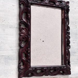 Puede incluir: Un espejo rectangular con un marco burdeos oscuro y ornamentado. El marco presenta un diseño tallado detallado con patrones de volutas. El espejo está sobre un fondo de madera blanca y desgastada, creando una estética vintage.