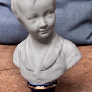 Könnte beinhalten: Eine weiße Porzellanbüste eines jungen Menschen mit detailliertem Gesicht und Schultern. Die Büste ist auf einem dunkelblauen Sockel mit goldenen Verzierungen montiert, was eine klassische Ästhetik erzeugt. Eine dekorative Skulptur.