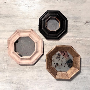 May include: Three octagonal mirrors with distressed frames. One is black, one is a pale pink, and the third is gold. The mirrors have a vintage, antique appearance, with aged glass and decorative frames. The mirrors are displayed on a light gray wooden surface.