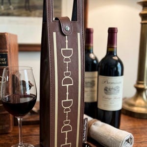 May include: A tall, brown, rectangular wine bottle carrier with a top handle and a snap closure. The front features a vertical gold-colored graphic of a horse bit design and the text "The Stallion Post Design" printed near the bottom edge.