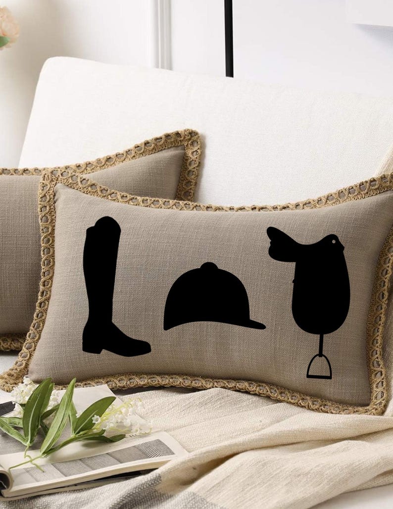 May include: Tan rectangular throw pillow with a black equestrian design featuring a riding boot, helmet, and saddle. The pillow has a decorative jute trim and is placed on a white surface.